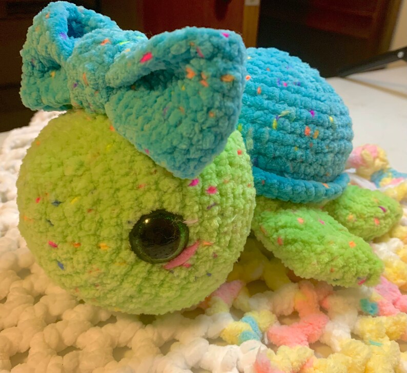 May include: A crocheted turtle toy with a blue bow and green and blue speckled yarn. The turtle has large, black eyes and a pink nose.