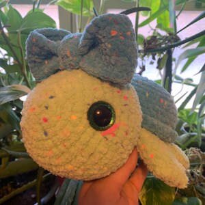 May include: A crocheted turtle plush toy with a blue bow and green and white speckled fur. The toy has large, sparkly green eyes with pink accents.