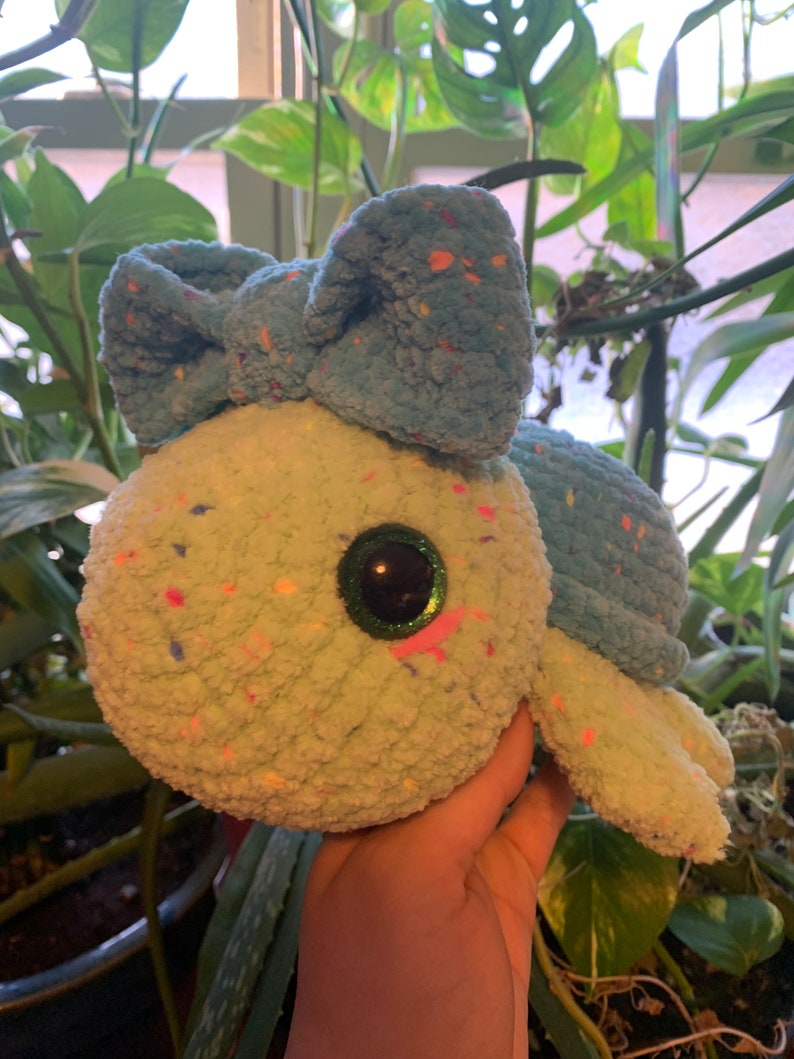 May include: A crocheted turtle plush toy with a blue bow and green and white speckled fur. The toy has large, sparkly green eyes with pink accents.