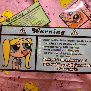May include: A blue and white warning sign with a cartoon girl with blonde pigtails and a pink bubble. The sign reads "Warning" and includes safety tips for children in cars. The text also says "Hot girl in drivers seat So buckle yo MF seatbelt & stay safe!"