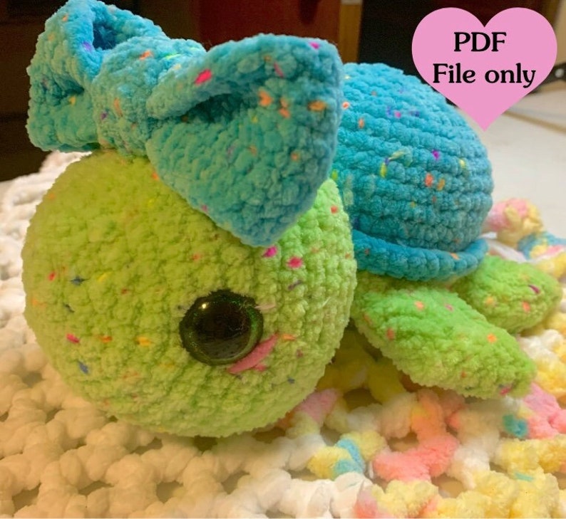 May include: A crocheted turtle amigurumi toy with a blue bow, green and blue speckled yarn, and large black eyes. The toy is sitting on a white and pastel-colored crocheted surface. PDF File only.