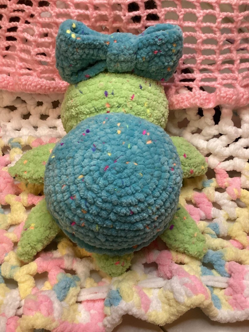 May include: A crocheted turtle toy with a blue shell and green body. The turtle has a blue bow on its head and is lying on a colorful crocheted blanket.