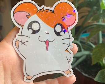Pode incluir: Um adesivo de hamster de desenho animado branco e laranja com um acabamento brilhante iridescente. O hamster está sorrindo e acenando com a pata direita.
