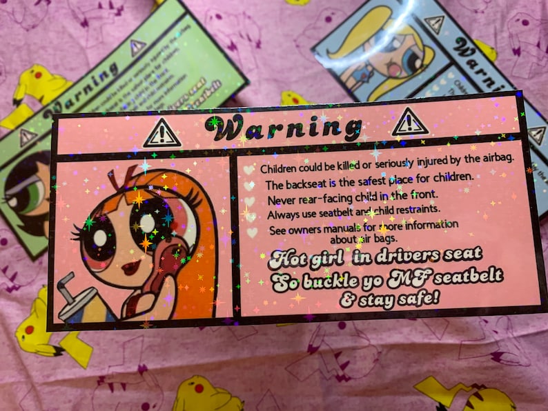 May include: A pink and white warning sign with a cartoon girl holding a drink. The sign reads "Warning" and includes safety tips for children in cars, such as "Children could be killed or seriously injured by the airbag." and "Hot girl in drivers seat So buckle yo MF seatbelt & stay safe!"