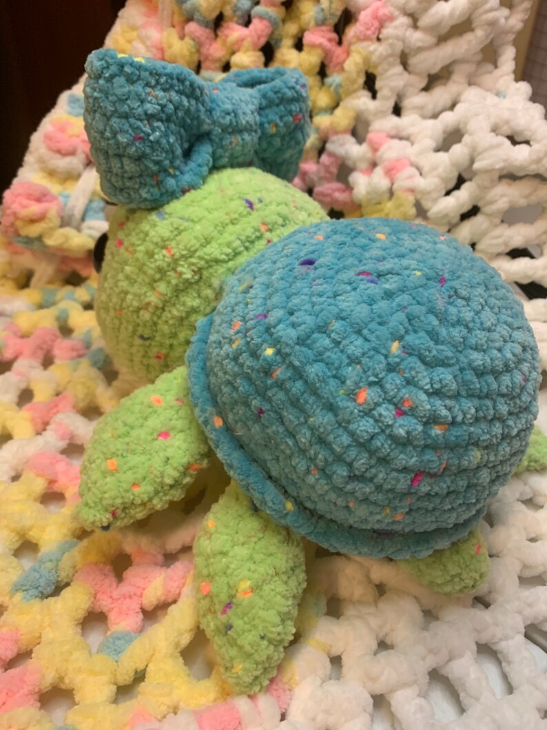 May include: A crocheted turtle toy with a green shell and blue head. The turtle has a blue bow on its head and is covered in colorful sprinkles.
