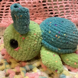 May include: A crocheted stuffed turtle toy with a green body, blue shell, and colorful speckles. The turtle has large, black eyes and a pink nose.
