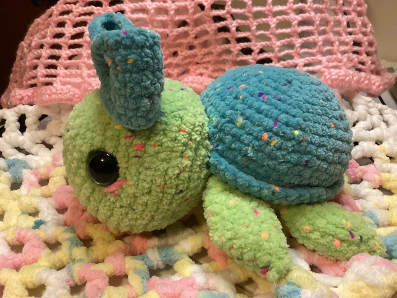 May include: A crocheted stuffed turtle toy with a green body, blue shell, and colorful speckles. The turtle has large, black eyes and a pink nose.