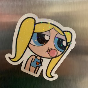 May include: A cartoon sticker of a girl with blonde hair, blue eyes, and a pink bubble. She is wearing a blue and red top. The sticker has a glittery finish.