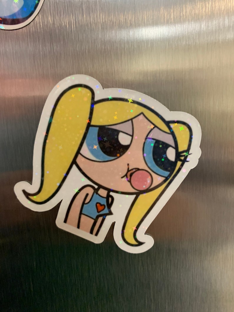May include: A cartoon sticker of a girl with blonde hair, blue eyes, and a pink bubble. She is wearing a blue and red top. The sticker has a glittery finish.