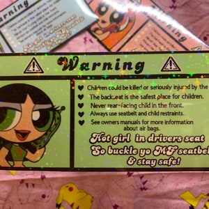 May include: A green and black warning sign with a cartoon girl holding a phone. The sign reads "Warning" and includes safety tips for children in cars, such as "Children could be killed or seriously injured by the airbag." and "Hot girl in drivers seat So buckle yo MF seatbelt & stay safe!"
