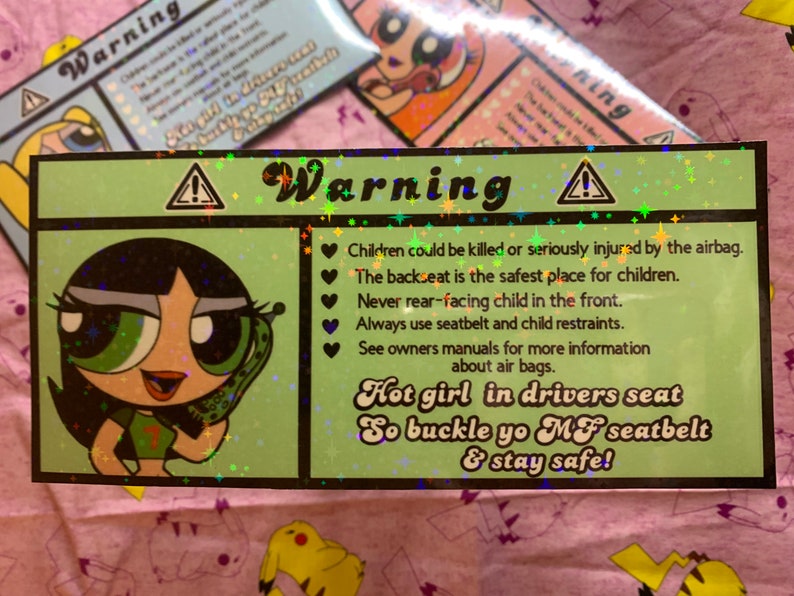 May include: A green and black warning sign with a cartoon girl holding a phone. The sign reads "Warning" and includes safety tips for children in cars, such as "Children could be killed or seriously injured by the airbag." and "Hot girl in drivers seat So buckle yo MF seatbelt & stay safe!"