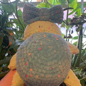 May include: A crocheted stuffed turtle toy with a light blue shell and a light green body. The shell has a speckled pattern with multi-colored flecks. The turtle has a large blue bow on its head.