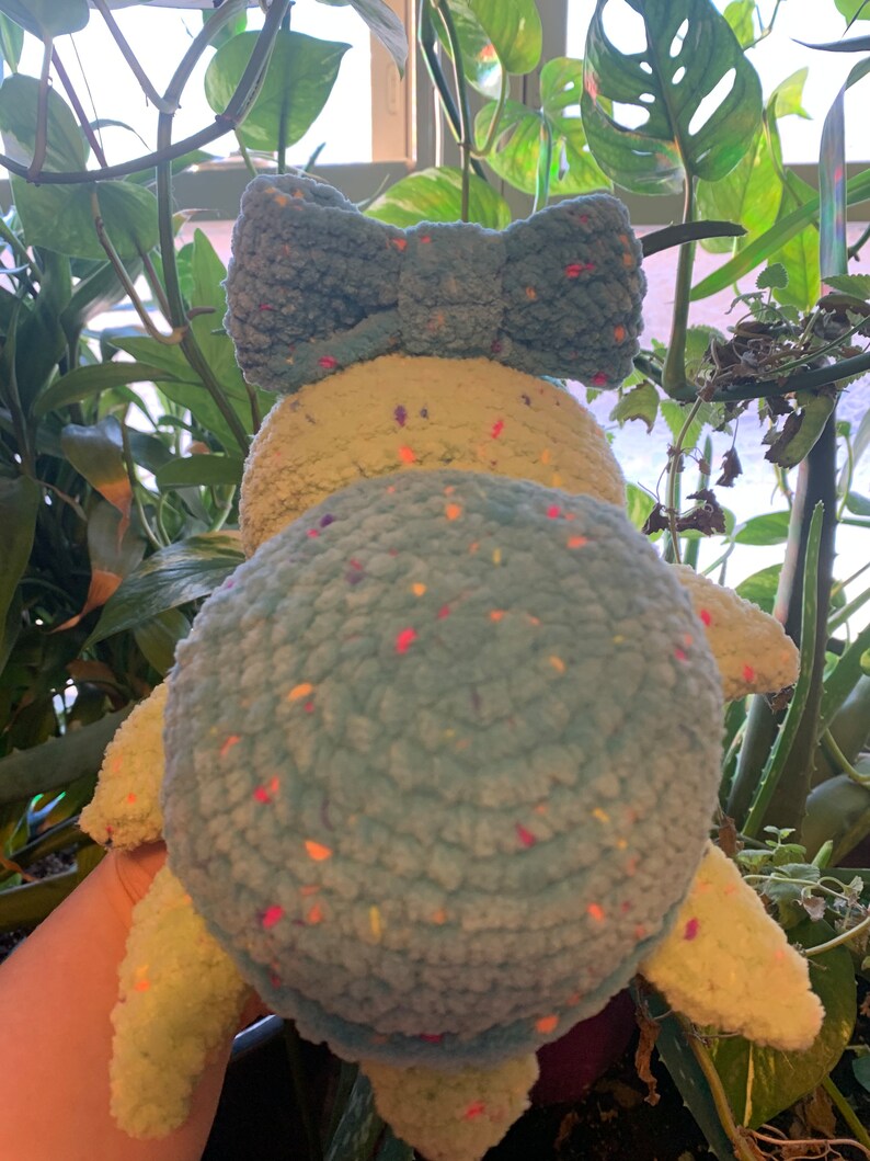 May include: A crocheted stuffed turtle toy with a light blue shell and a light green body. The shell has a speckled pattern with multi-colored flecks. The turtle has a large blue bow on its head.