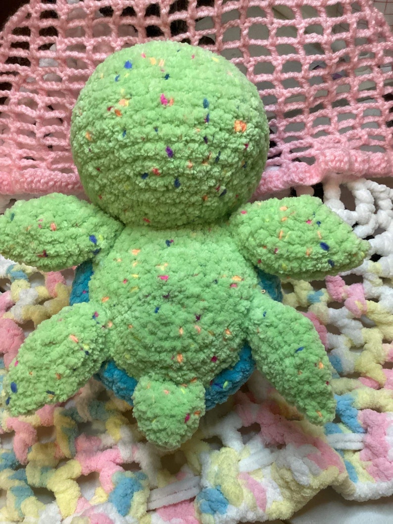 May include: A green crocheted turtle stuffed animal with colorful flecks throughout the fabric. The turtle is sitting on a multi-colored crocheted blanket.