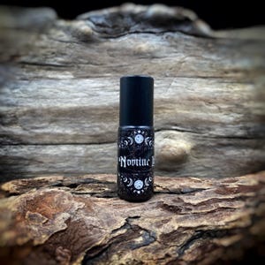 May include: A black rollerball bottle with a black cap. The label features a white moon and stars design with the word "Nomine I" in white script. The bottle is displayed on a piece of weathered wood.