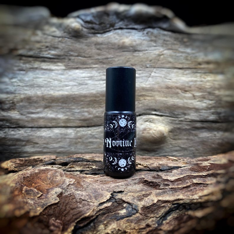 May include: A black rollerball bottle with a black cap. The label features a white moon and stars design with the word "Nomine I" in white script. The bottle is displayed on a piece of weathered wood.