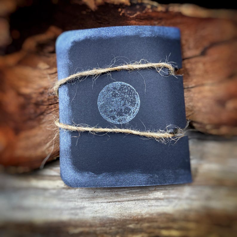 May include: A dark blue, rectangular notebook with a detailed moon design in the center. The notebook is wrapped with natural twine. The background is a blurred wooden surface.