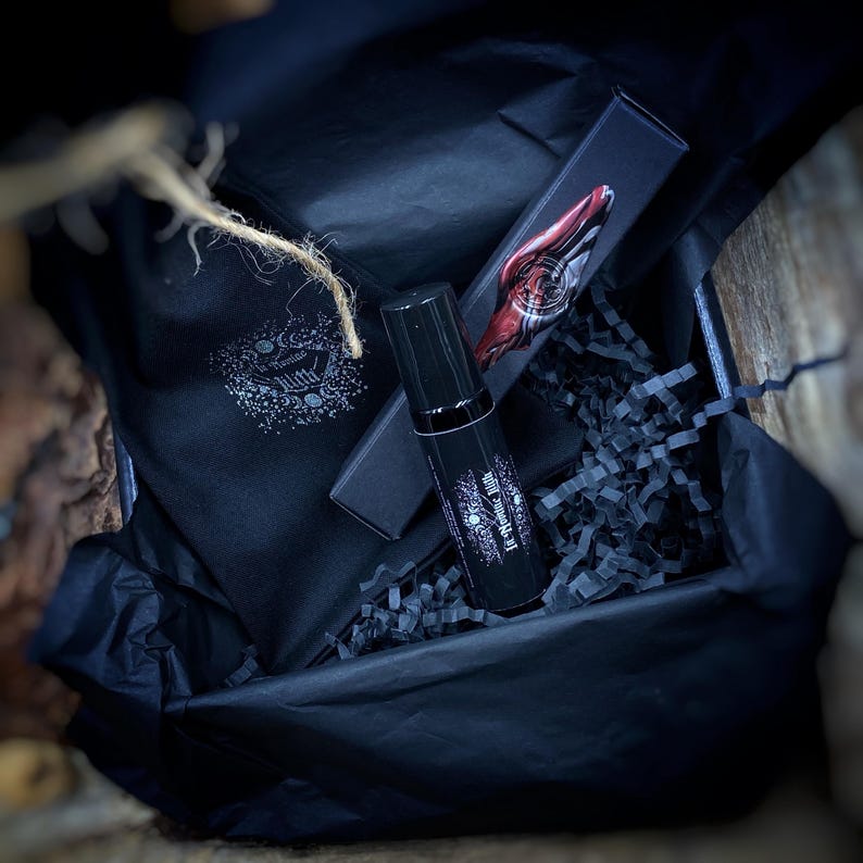 May include: A black gift box with black tissue paper, a black bottle, and a black box. The bottle has white text. A black cloth bag with a white design is also in the box. The box is open, revealing the contents.