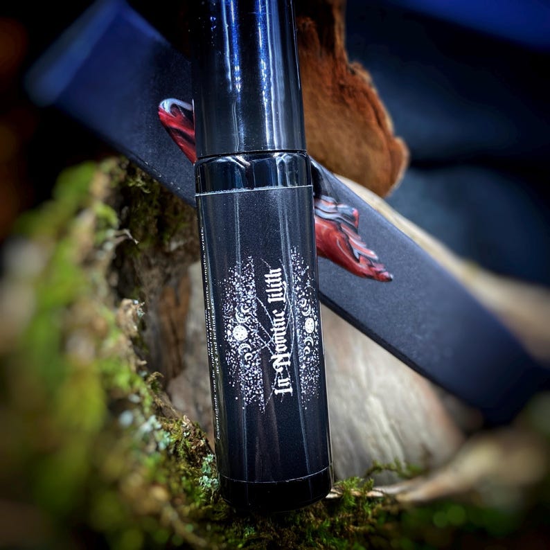 May include: A black roll-on perfume bottle with the text "In Nomine Lilith" in white script. The bottle is on a mossy surface, with a dark box featuring a red hand design in the background.