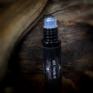 May include: A black roll-on bottle with a clear rollerball and cap. The bottle has a label with white text that reads "In Nomine Lilith" and decorative elements. The bottle is cylindrical and appears to be made of glass.