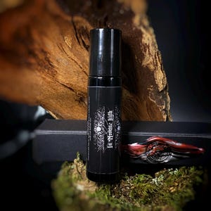 May include: A black roll-on bottle with white text and a decorative design. The bottle is labeled "In Nomine Lilith". A red and silver decorative object is next to the bottle. The background includes a piece of wood and moss.