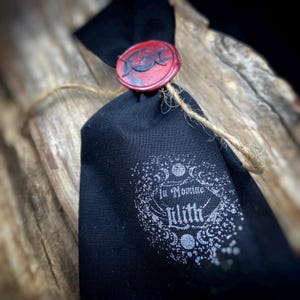 May include: A black fabric bag tied with twine, sealed with a red wax stamp featuring a crescent moon design. The bag has the words "In Nomine Lilith" printed in white, surrounded by a decorative border.