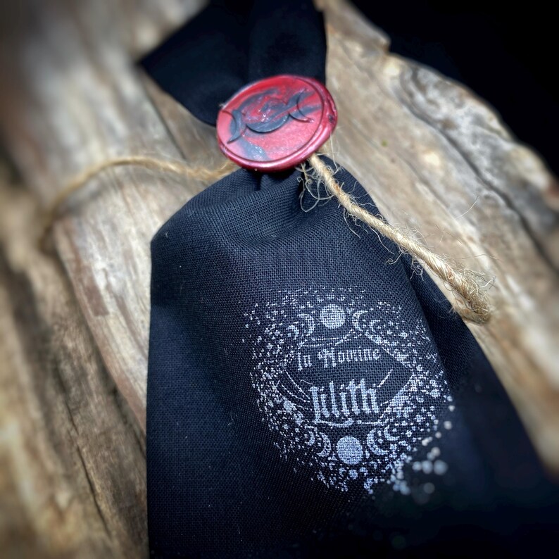 May include: A black fabric bag tied with twine, sealed with a red wax stamp featuring a crescent moon design. The bag has the words "In Nomine Lilith" printed in white, surrounded by a decorative border.