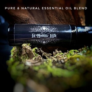 May include: A black essential oil blend bottle with the text "In Nomine Lilith" and "Pure & Natural Essential Oil Blend". The bottle is set on a bed of green moss and wood.