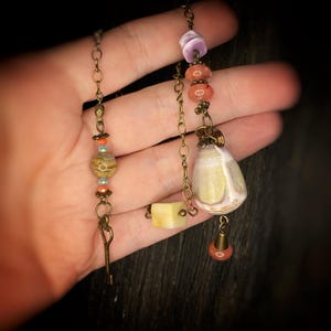 May include: A long, multi-strand necklace with a variety of beads, including a large, white shell pendant, a small, square, yellow stone, and several smaller, pink and orange beads. The necklace is made with a brass chain and has a key charm.