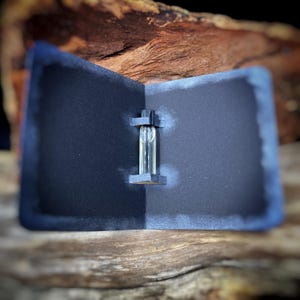 May include: A small, clear glass vial containing a liquid, held in place by a dark gray, folded card. The card is set against a backdrop of wood and a dark background. The vial has a black cap.