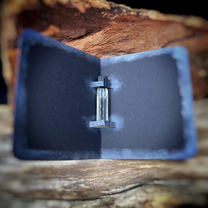 May include: A small, clear glass vial containing a liquid, held in place by a dark gray, folded card. The card is set against a backdrop of wood and a dark background. The vial has a black cap.