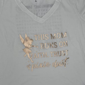 May include: White t-shirt with a gold glitter design featuring a fairy and the text "This Mom Runs On Faith, Trust & Pixie Dust."