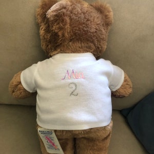 May include: A brown teddy bear wearing a white t-shirt with the name "Mia" and the number "2" embroidered on the back. The bear has a Build-A-Bear Workshop tag attached to its leg.