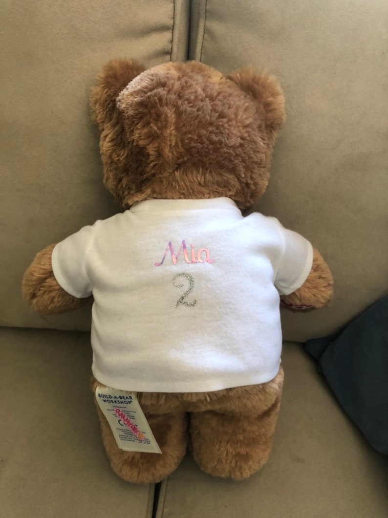 May include: A brown teddy bear wearing a white t-shirt with the name "Mia" and the number "2" embroidered on the back. The bear has a Build-A-Bear Workshop tag attached to its leg.