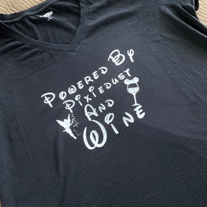 May include: Black t-shirt with white text that reads "Powered by Pixie Dust and Wine". The text is stylized with a fairy, a wine glass with Mickey Mouse ears, and a Disney-inspired font.