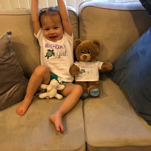 May include: A young child wearing a white shirt with the text "Birthday girl" and a green turtle graphic sits on a beige couch with a brown teddy bear wearing a matching shirt. The teddy bear has blue and white polka dot feet.