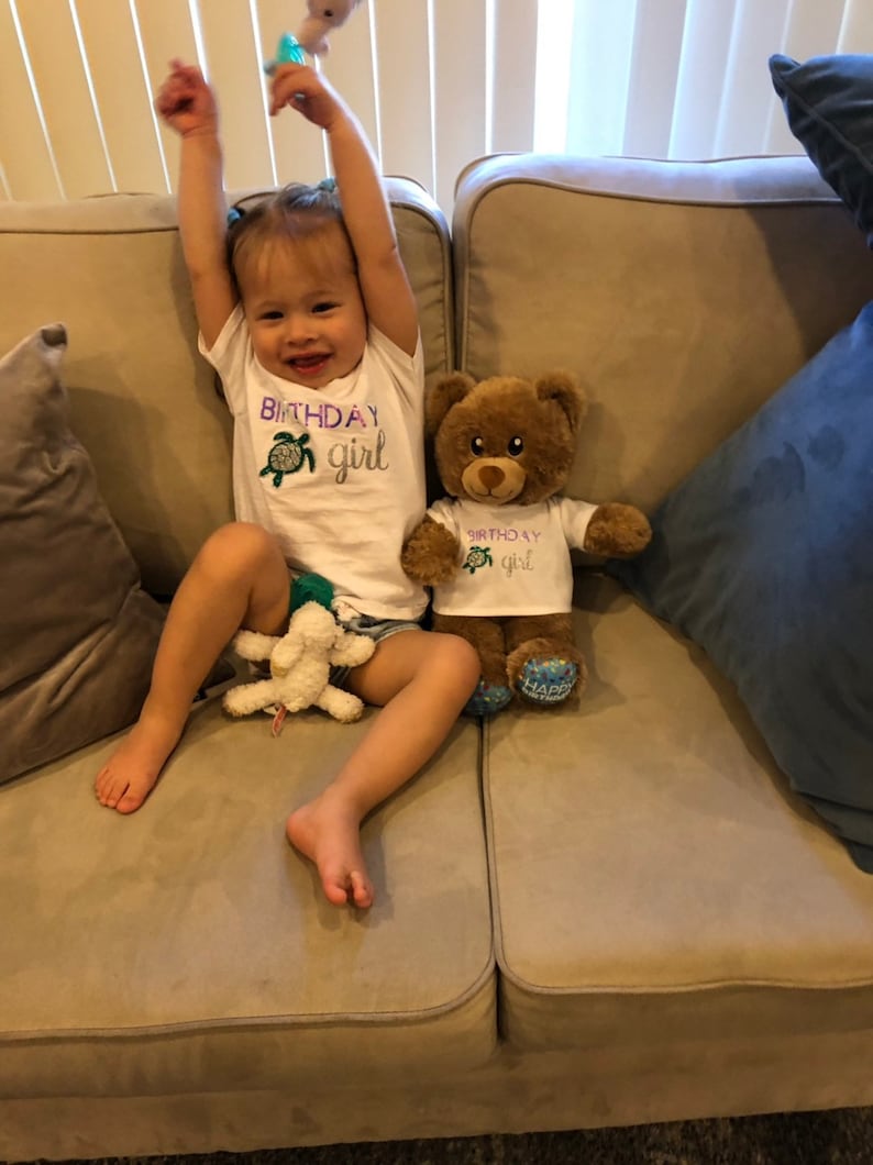 May include: A young child wearing a white shirt with the text "Birthday girl" and a green turtle graphic sits on a beige couch with a brown teddy bear wearing a matching shirt. The teddy bear has blue and white polka dot feet.