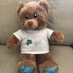 May include: A brown teddy bear wearing a white t-shirt with the text "Birthday girl" and a green sea turtle design. The bear has blue feet with a colorful "Happy Birthday" design.