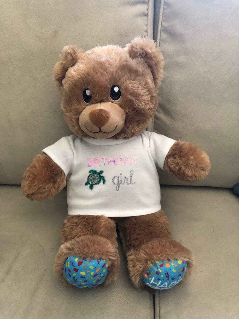 May include: A brown teddy bear wearing a white t-shirt with the text "Birthday girl" and a green sea turtle design. The bear has blue feet with a colorful "Happy Birthday" design.