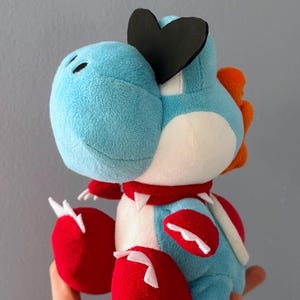Könnte beinhalten: Ein Plüschtier eines blauen, dinosaurierartigen Charakters mit roten Füßen und einem roten Sattel. Das Spielzeug hat ein schwarzes herzförmiges Element auf dem Kopf und einen roten Kragen mit weißen Stacheln. Das Spielzeug wird in einer Hand gehalten.