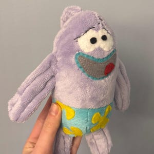 Puede incluir: Un peluche del personaje Patricio Estrella de la serie animada Bob Esponja. El juguete es de color lavanda con una cara sonriente, un bañador azul y amarillo y brazos extendidos.