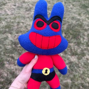 May include: A plush toy character with a blue head and ears, red eyes and a wide red smile. The body is red with blue limbs. It wears a black belt with a gold buckle. The toy is designed to resemble a cartoon character.