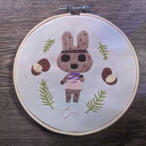 Könnte beinhalten: Stickbild mit Stickerei, das ein braunes Kaninchen mit einem lila und rosa Kleid zeigt, umgeben von grünen Blättern und braunen Nüssen. Der Name "Coco" ist unter dem Charakter gestickt.