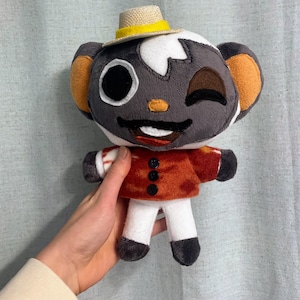 Pode incluir: Um brinquedo de pelúcia de um animal cinza e branco vestindo um chapéu amarelo e marrom e uma camisa listrada vermelha e branca. O brinquedo tem um olho piscando e um grande sorriso.