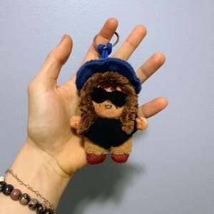 May include: A plush keychain featuring a small figure with brown hair, wearing a dark blue hat, black sunglasses, and a black outfit. The figure has tan skin, a small smile, and red shoes. A blue cord attaches the plush to a silver metal keyring.