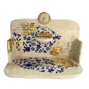 May include: A cream-colored ceramic shelf with a flat base and a back panel featuring blue floral patterns, gold leaf accents, and a small circular emblem at the top center. The structure includes a small protruding ledge and textured details.