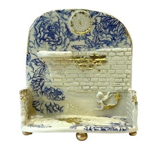 May include: A decorative ceramic shelf with a blue and white floral design. The shelf has a brick wall background with gold accents and a small gold figure. The base has three gold feet.