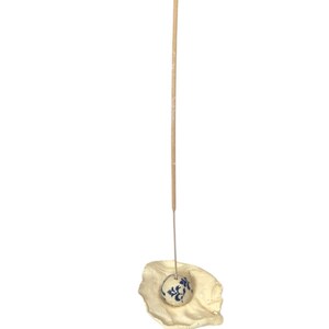 May include: A beige incense holder with a small, round, blue and white floral patterned ceramic ball. A thin, tan incense stick is inserted into the ball, and the holder has a textured, organic shape.
