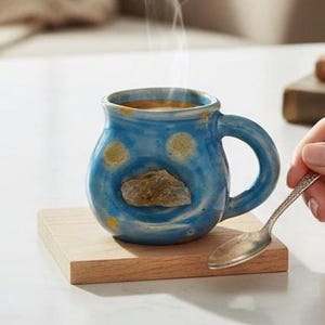 May include: A blue ceramic mug with a unique design, featuring a stone accent and yellow polka dots. The mug sits on a wooden coaster, with steam rising from the contents. A silver spoon is held nearby.