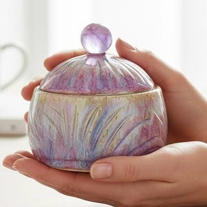 May include: A small, round ceramic jar with a lid, held in cupped hands. The jar is a blend of lavender, pink, and yellow hues, with a carved leaf design. The lid features a small, round, purple knob.
